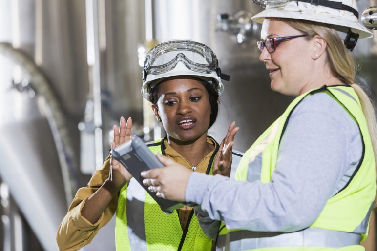 Two multiracial women working together in a manufacturing plant, standing in front of steel storage tanks.  They are wearing white hardhats with safety goggles, and yellow reflective vests, looking at the screen of a digital tablet.  The African American woman is talking and gesturing.